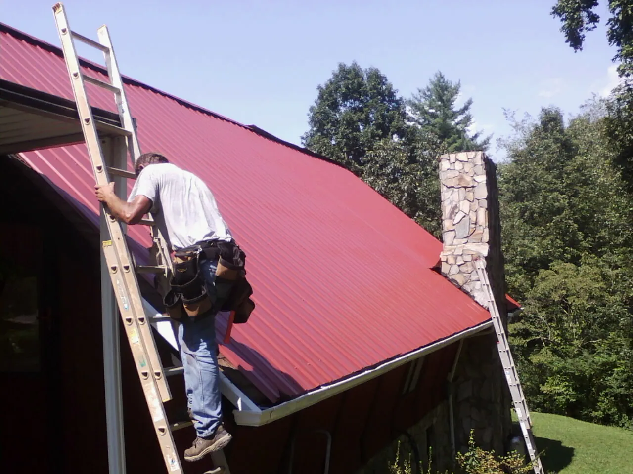 Roofer on a ladder beside a red metal roof and stone chimney — Soffit & Fascia Repair by Roofing Brothers in Medusa